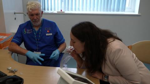 A woman with long dark hair and wearing a light pink jacket leans forward towards a desk.  She is blowing into a tube on a clear plastic bag which is attached to a small white machine with a built in monitor.  To her left is a nurse watching her progress. He is wearing royal blue scrubs and blue latex gloves and has white hair and a short white beard.  