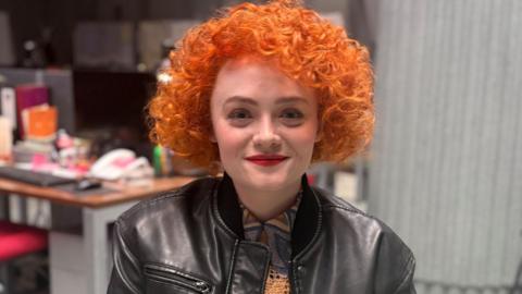 Leah Byrne, who plays DC Rose Dickson, sitting on a seat inside the building. A desk and chair can be seen over her right shoulder and a grey pillar is behind her left shoulder. She has short, bright orange curly hair and is wearing a black leather jacket over an orange top