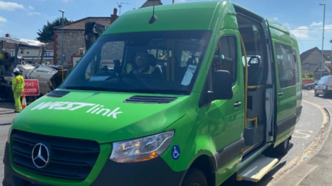 A green minibus is parked on a street. The side door is open. The bus has Westlink in white writing on the bonnet. 