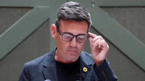 Andy Burnham looks down, holding the side of his glasses. He wears a blue jacket, blue top and a yellow badge with a bee symbol on it.