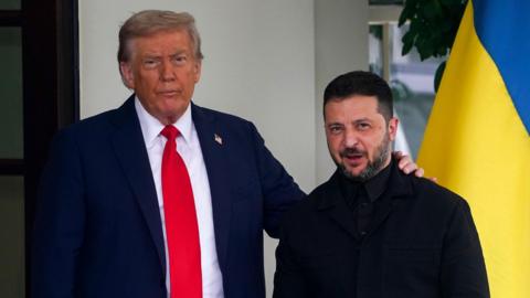 Donald Trump, wearing a blue suit and red tie, places a hand on the shoulder of Volodymyr Zelensky, dressed in black, with a Ukrainian flag visible behind them