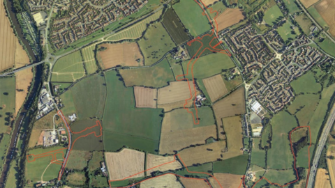 An aerial view of a number of large fields, with existing housing developments to the right and top of the view. A road runs across the bottom left of the image, and there are red lines on the map showing where new roads would go