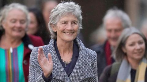 Catherine Connolly has short grey hair. She is wearing a navy suit with a blue and grey overcoat which has large lapes. She is wearing a silver necklace, is smiling and waving.
