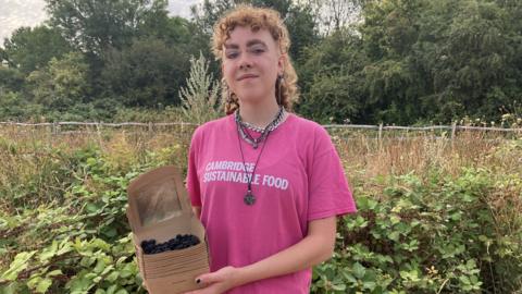 Alex Vardill wearing a pink T-shirt, smiling straight at the camera and holding a box of blackberries.