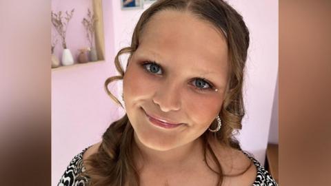 A young girl aged around 12. She has curly hair, earrings and she is wearing a black and white patterned top. She is smiling into the camera.