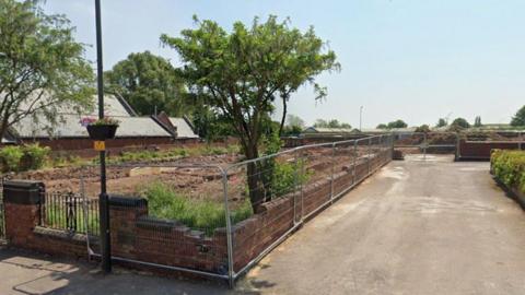 A site with uneven dirt on it, surrounded by a low brick wall and silver wire fencing.