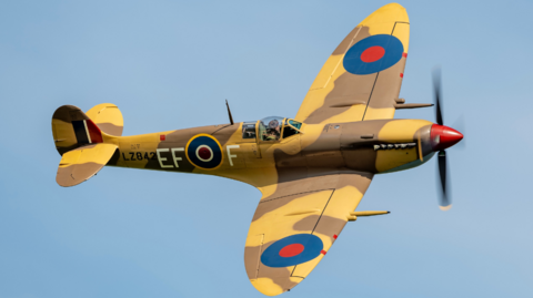 A Mark 9 Spitfire - designated callsign LZ842 - painted in yellow and brown desert camouflage. There are blue and red circles on top of each wing, which are the emblem of the Royal Air Force. The plane is flying against a blue sky, and its propeller is turning around a red nose cone.