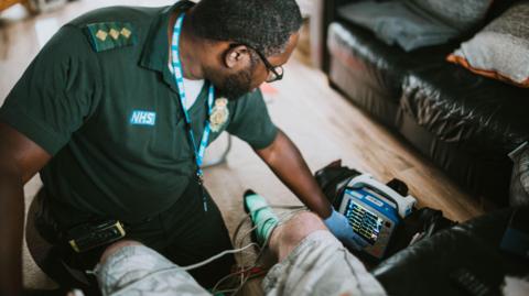 A paramedic, wearing a green polo shirt and glasses, assesses a patient at home.