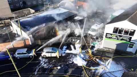 Four firefighters are seen from above spraying water on a smoldering industrial building. 