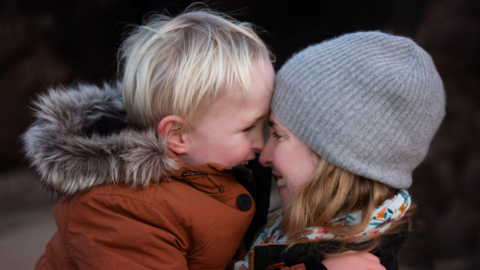 Close up of a woman a holding a young boy. The smiling at each other and are nose-to-nose. The woman is wearing a beanie hat and a floral scarf. The young boy is wearing a zip up coat with a fur-lined hood.