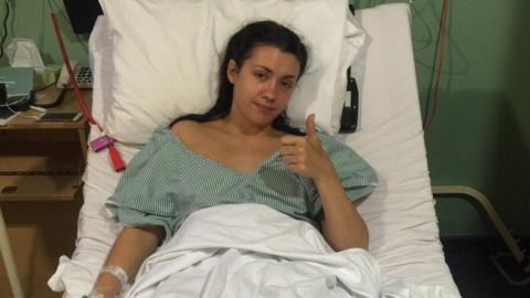 A woman lies in a hospital bed. Her legs are covered in a blanket. She has a cannula in one arm. She shows a thumbs up on her other hand.