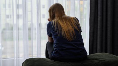 A young woman with long brown hair sits on the edge of a bed and looks out of a window 