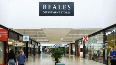 A line of shops along both sides with people walking - planters stand in the centre and an escalator is at the rear.