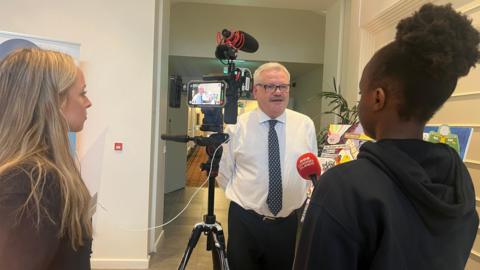 Two young girls stand in front of a camera interview a politican. The girl on the right is holding a BBC Channel Islands mic and is facing the poitician. The other girls is to the left and standing behind an iPhone camera watching the recording.