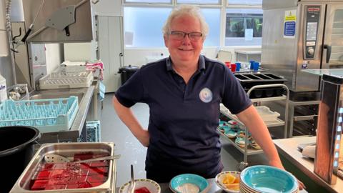 A dinner lady with short grey hair standing in a school kitchen 
