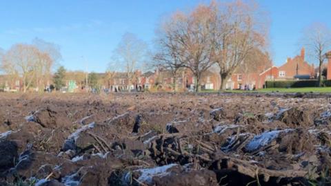 A churned-up muddy field in Central Park, Boston. In the background are bare trees and red-brick houses.