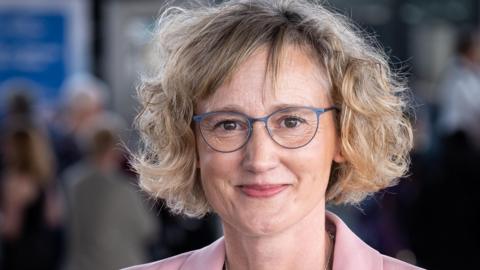 Welsh language Commissioner Efa Gruffudd Jones. She has a blonde short curly bob with a side fringe. She wears grey glasses and her pink collar can be seen in the bottom of the frame. She has brown eyes and smiles at the camera. 