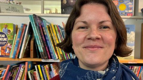 A woman smiles and looks directly at the camera. She is standing in front of a number of bookshelves filled with colourful children's books. She has brown hair, brown eyes and is wearing a red cardigan and blue patterned scarf.