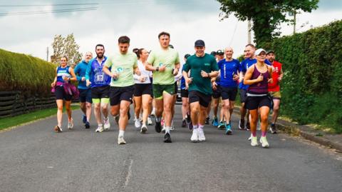 A large group of people are running on a road towards the camera. There's about 12 to 20 people in the ground. At the front are two young men in identical running gear - a light-green top, dark green shorts, trainers and white socks. The road is lined with hedges and in the background there's a cloudy sky.
