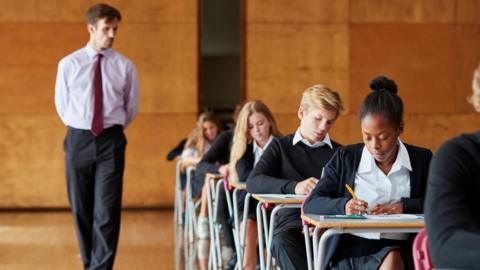 Students in a large hall seated at individual desks sitting an exam with a male teacher stood beside them