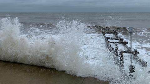 A large wave crashed onto the beach at Walcott, Norfolk, on a grey and cloudy day. To the left of the picture is a wooden flood defence used to break up the waves.