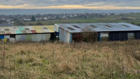Corrugated iron farm buildings on empty green land overlooking hills with houses in the distance