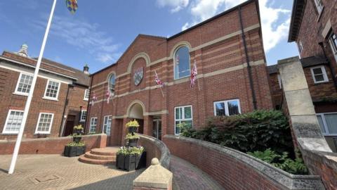 Exterior of South Kesteven council offices a two-storey brick building with the council's coat of arms over the main door and four union flags mounted on poles on the front of the building