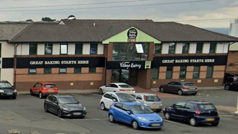 The exterior of The Village Bakery. In front of the building is a car park with a number of cars parked in it