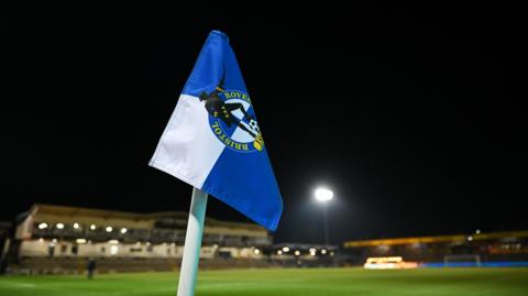 Bristol Rovers FC flag with a stand at the Memorial Stadium in the distance. The flag is blue and white with the club's emblem in the middle.