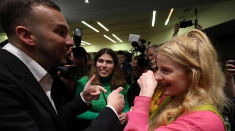 Green Party leader Zack Polanski speaks to Green Party MP Hannah Spencer who smiles as photographers take pictures around them. A party worker stands in the background.