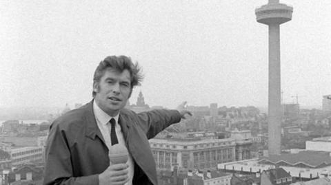 Nationwide reporter Bob Langley holding a microphone and pointing at the Liverpool Beacon, a tall tower with observation platform in Liverpool city centre.