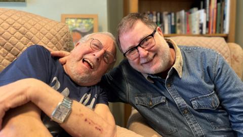 Llun o Danny Mendoza, ar y dde, gyda'i dad, sydd ar y chwith. Mae Danny'n gwisgo crys denim glas a sbectol ddu. Mae ei dad yn gwisgo crys-t glas tywyll, oriawr a sbectol grwn. Mae'r ddau'n gwenu'n tra'n eistedd mewn cadeiriau esmwyth.