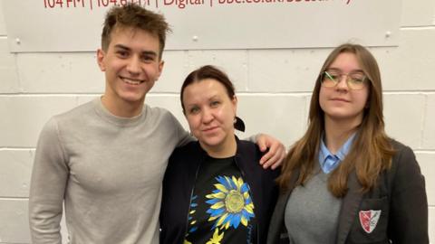 A mum and her teenage son and daughter stood in front of a white wall. All three are looking directly at the camera. 