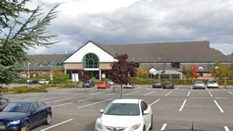 Image shows a large car park with a number of cars parked in it. 
In the distance is a large double storey building - which is the hotel.
It has an arched window at the reception area. 