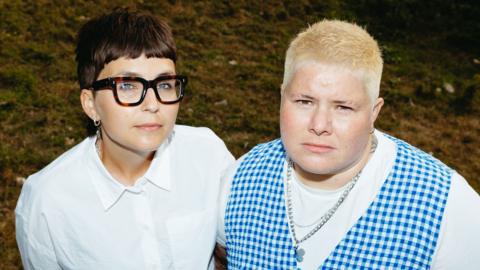 a mid-length image of the band arxx standing in a field. clara is on the left, and has short brunette hair wearing thick-rimmed tortoiseshell glasses, a silver chain and a white shirt. hanni is on the right. they have short platinum blonde hair, and are wearing a silver chain, a white t-shirt and a blue gingham waistcoat