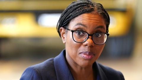A picture of Kemi Badenoch from the shoulders up. She is wearing a dark blue suit jacket and black-rimmed glasses. Her hair, which is in braids, is tied back. She is looking to the left and the background is blurred.