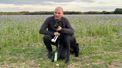BBC correspondent Sean Dilley pictured in a field on a bright day, crouched down with his arms around his black guide dog Shawn