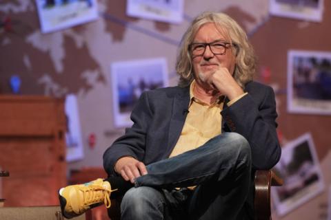 James May has long wavy grey hair and glasses. He is wearing jeans, a yellow shirt and a navy blazer.