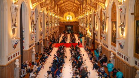 Six rows of people sit facing white table cloths seated for the Iftar event