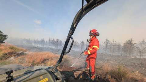 A firefighter in bright red protective clothing stands in a smoke-filled heathland, spraying water onto scorched ground after a fire. The scene is viewed from inside an off-road vehicle, with part of the vehicle frame visible in the foreground. Burnt vegetation and a line of smoke-shrouded trees stretch across the background under a pale blue sky.