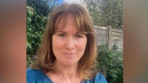 A woman with shoulder-length hair wearing a blue top. She is stood outdoors with greenery and a fence in the background of the selfie.