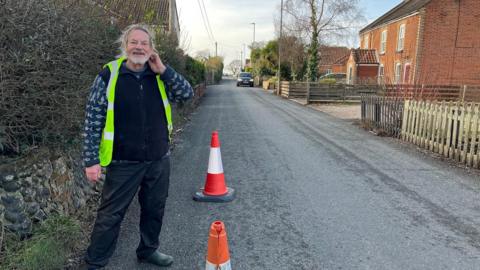 Berni Marfleet standing on Aylsham Road in North Walsham wearing a yellow high-vis jacket. He has long grey hair tied in a ponytail. He is wearing a blue jumper with a grey pattern on it and black trousers