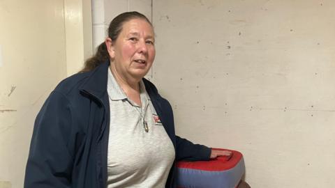A woman standing indoors beside stacked training pads against a worn, marked wall.
