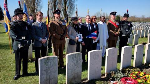 Eight people, including civilians in smart clothes, a priest in white robes s and men in military uniforms stand in a line in front of a row of white headstones. Behind them are military personnel holding flags. Rows of gravestones can be seen beyond. The sky is blue.