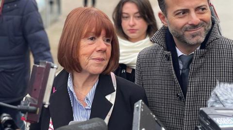 Gisèle Pelicot is surrounding by members of the press carrying cameras. She is walking with her head held high with a faint smile. She has a red bob and is wearing a black coat. She is flanked by her legal team.