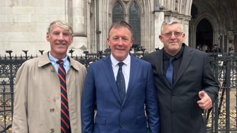 Left to right - Rob Sheridan in a blue shirt, blue and orange striped tie and a beige raincoat, Neil Stevens in a white shirt, navy blue tie and suit jacket, Tony Stevens in a black shirt with a navy blue tie and a grey suit jacket, all standing in front of the Royal Courts of Justice.