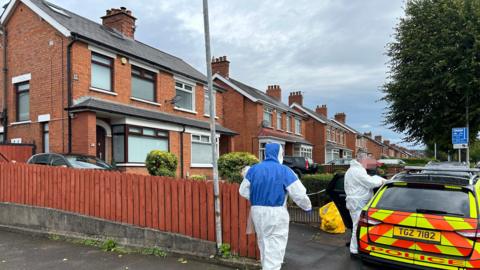 Forensic officer in foreground walking towards a semi-detached house. Police car outside house with blurred faces of two people standing beside it.