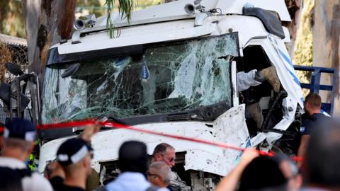 A mangled truck with a smashed windscreen surrounded by emergency services