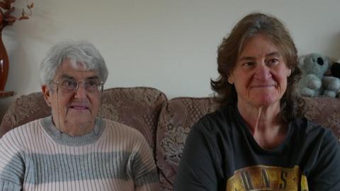 An image of Theresa (on the left) and Sharon Waring (on the right).  Theresa is wearing a pink and grey stripy jumper, while Sharon has on a black Guns N' Roses band t-shirt.  They are both sitting on a pink floral settee.  There is a grey stuffed teddy bear behind Sharon.