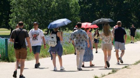 people walking on a pathway through a park. Some are holding umbrellas to shade them from the hot weather